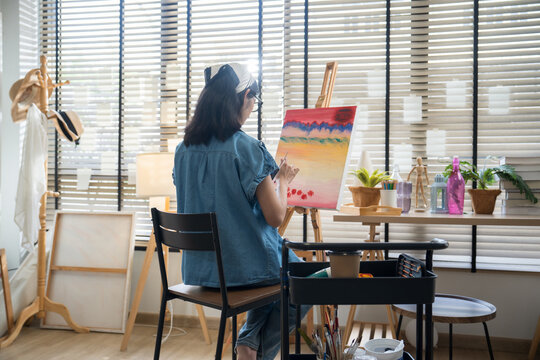 Young creative female artist sitting on a chair, painting on canvas with watercolors in a bright home art studio.
