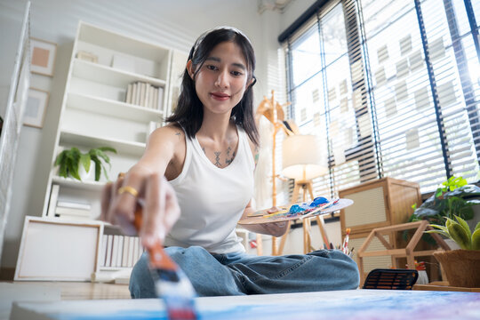 Creative tattooed young woman sitting cross-legged on the floor, mixing paint on a color palette to paint on paper in a bright home art studio.