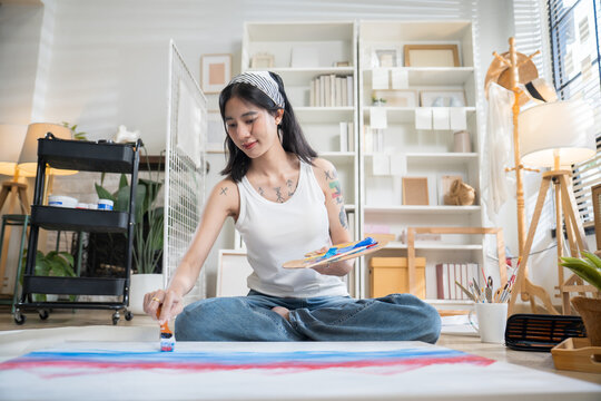 Creative tattooed young woman sitting cross-legged on the floor, mixing paint on a color palette to paint on paper in a bright home art studio.