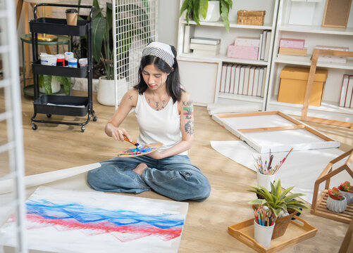 Creative tattooed young woman sitting cross-legged on the floor, mixing paint on a color palette to paint on paper in a bright home art studio.
