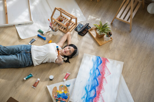 Top view of creative tattooed female artist lying on floor, holding paintbrush with passion and dreaming near her colorful painting.