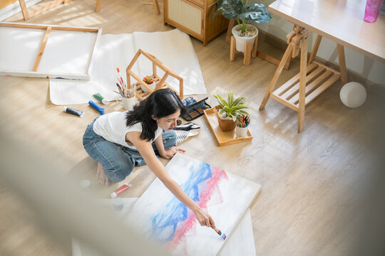 Top view creative young woman sitting cross-legged on the floor, mixing paint on a color palette to paint on paper in a bright home art studio.