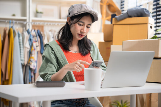 Female small business owner wearing a hat, sitting at her desk and checking orders on her smartphone with a laptop nearby.
