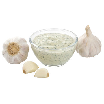 Clear glass bowl of creamy garlic sauce with fresh minced herbs, accompanied by two whole garlic bulbs and two peeled cloves, isolated on a white background