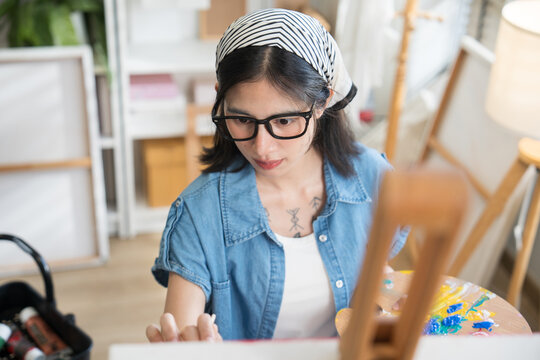Young creative female artist sitting on a chair, painting on canvas with watercolors in a bright home art studio.