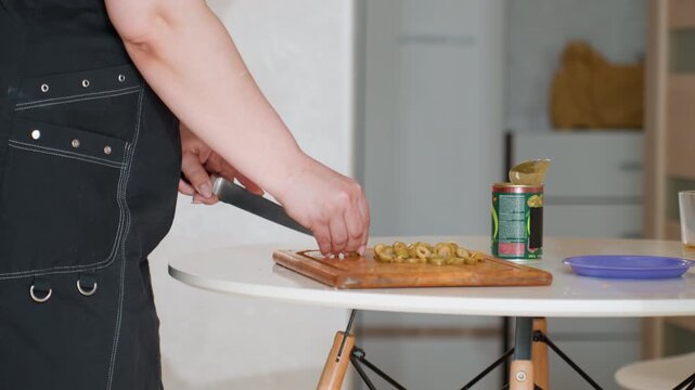 Close-up of hand using knife to slice fresh vegetables on wooden board beside round white table, showing active food preparation, neat kitchen setting, natural light, and casual home cooking