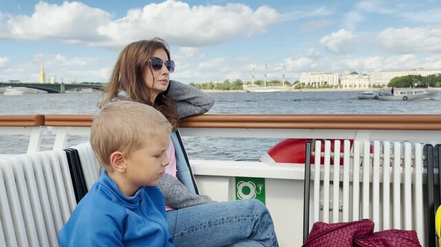 A woman and boy rest thoughtfully on a boat deck in Saint Petersburg. They gaze across the calm river past Trinity Bridge. This peaceful boat ride brings a quiet, serene feeling.