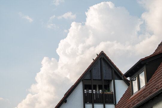 Sich auft&uuml;rmende wei&szlig;e Wolkengebilde vor blauem Himmel &uuml;ber Hausgiebel mit Rabenv&ouml;geln und Ziegeldach mit Fenster in Dachgaube in Dachgeschosswohnung in Neckartenzlingen.