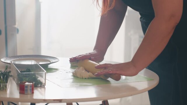 Close mid-angle view of person rolling and flattening two round dough portions on floured small round table, using rolling pin with steady hands under soft natural window light, with baking, rolling