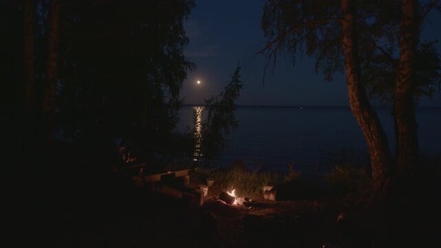 Bonfire burns on the campsite surrounded by trees and with the Moon rising on the background