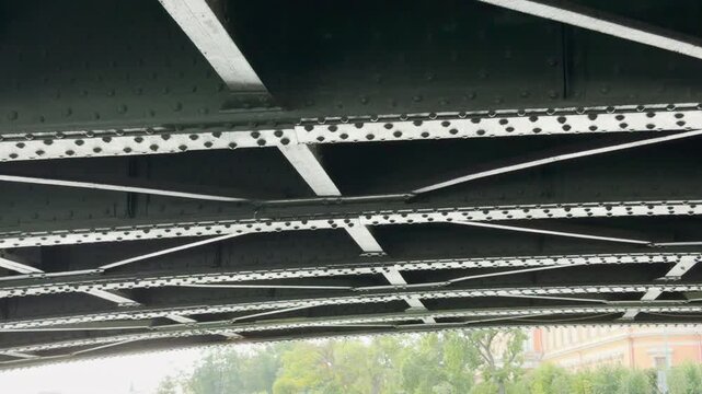 A historic metal bridge arches over the calm canal in Saint Petersburg. Heavy steel beams inspire a peaceful sense of awe. Bright trees line the sunny embankment ahead.