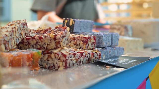 A market stall features colorful sweets arranged neatly for sale. These treats, made with nuts and fruits, attract attention from visitors of different ages and backgrounds