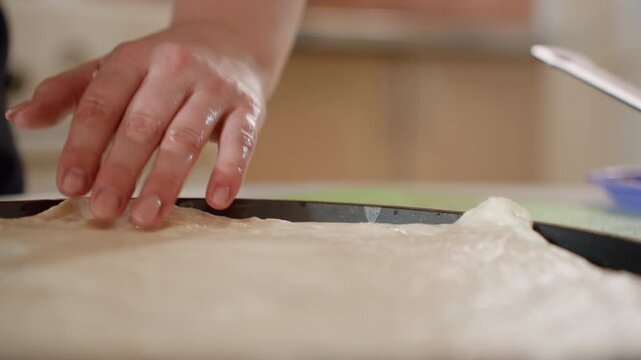Close-up shows adult hands pressing, stretching, and smoothing soft pizza dough on kitchen countertop, with light flour dust, steady shaping motion, and tactile surface detail during homemade