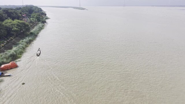 Drone aerial footage showing a wooden boat approaching the camera across the wide Padma River, leaving a long wake behind it, with green riverbank vegetation and transmission towers.
