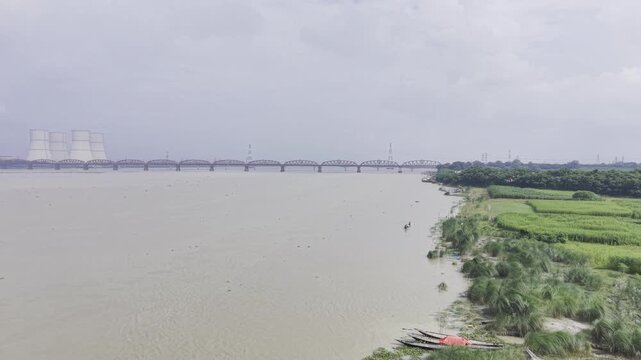 Aerial footage of a wooden boat speeding toward a stationary drone on the Padma River, with both riverbanks, Hardinge Bridge, Lalon Shah Bridge and cooling towers visible in the distance.