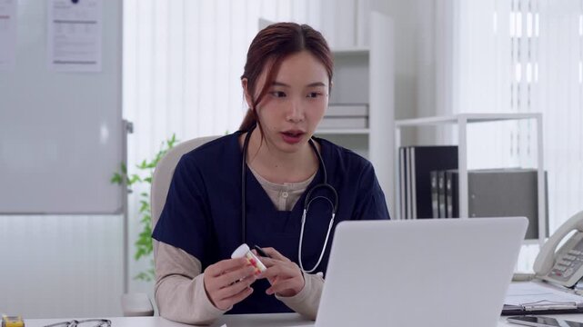 Young Asian female doctor consulting patient via telemedicine holding medication bottle during online appointment at modern clinic desk