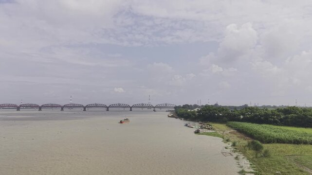 Forward-moving drone footage over the Padma River, with Hardinge Bridge and Rooppur Nuclear Power Plant cooling towers far away, while cargo boats approach the camera on the water.
