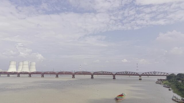 Aerial footage over the Padma River showing distant Hardinge Bridge and Rooppur Nuclear Power Plant cooling towers, while cargo boats approach the camera under a blue sky with clouds.