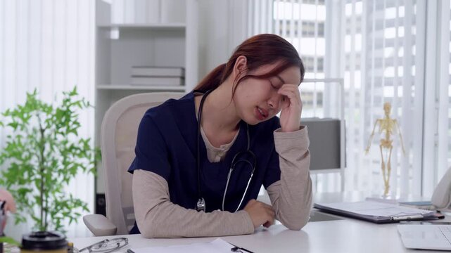 Young Asian female doctor experiencing headache and stress while working at medical office desk with city view