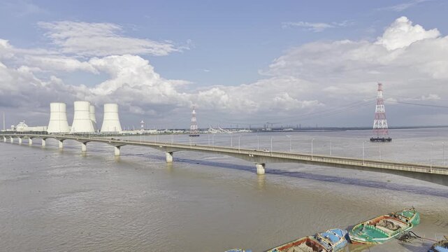 Drone aerial footage showing Lalon Shah Bridge across the Padma River, Rooppur Nuclear Power Plant cooling towers, transmission towers, cargo boats, blue sky and scattered white clouds in Bangladesh.