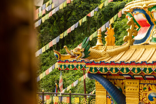 View of the ornate golden roof of a Buddhist temple adorned with a dragon sculpture and colorful prayer flags against a backdrop of lush trees in Pokhara, Gandaki Province, Nepal.