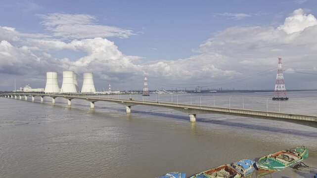 Aerial drone footage of Lalon Shah Bridge crossing the Padma River, with Rooppur Nuclear Power Plant cooling towers, cargo boats, blue sky and white clouds visible across the river in Bangladesh.