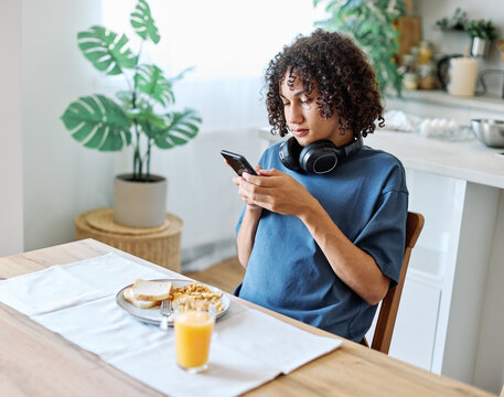 A teen sits in a bright modern kitchen, focused on a smartphone while eating breakfast with headphones around the neck. Casual morning routine captured with orange juice, toast and cereal on the table