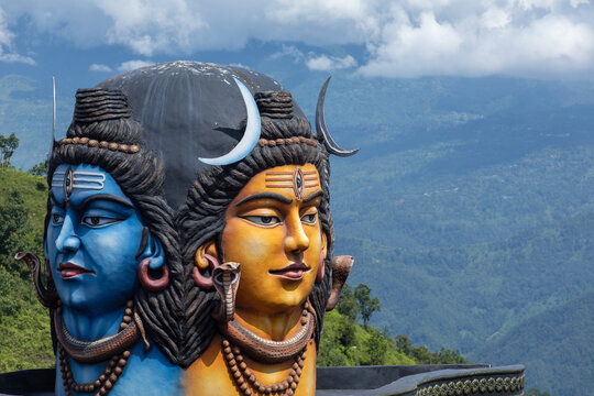 View of the dual-faced Shiva statue with blue and yellow skin, adorned with a crescent moon and snakes, set against lush green hills and mountains in Pokhara, Gandaki Province, Nepal.