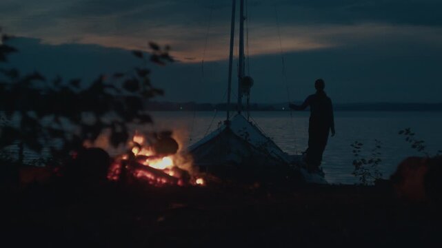 Sailing and camping in the wild. Woman sailor walks on the wild area with sailing boat on the background and bonfire on the foreground