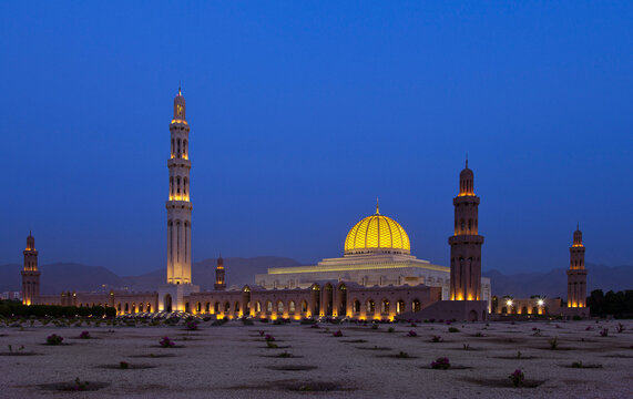 View of Sultan Qaboos Grand Mosque illuminated at twilight with its golden dome and minarets glowing against a deep blue sky in Muscat, Muscat Governorate, Oman.