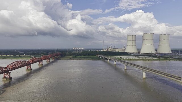 Aerial view of the wide Padma River between Hardinge Bridge and Lalon Shah Bridge, with Rooppur Nuclear Power Plant cooling towers, Green City Rooppur buildings, blue sky and scattered clouds visible.