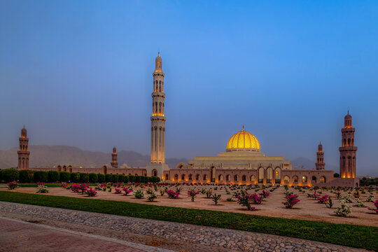 View of the illuminated Sultan Qaboos Grand Mosque with its golden dome and minaret rising above landscaped gardens in Muscat, Muscat Governorate, Oman.