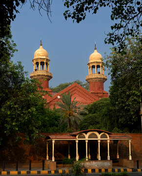View of the historic red brick architecture of the Lahore High Court with its distinctive domes framed by lush green trees and a clear blue sky in Lahore, Punjab, Pakistan.