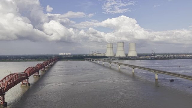 Aerial view across the Padma River between Hardinge Bridge and Lalon Shah Bridge, showing Rooppur Nuclear Power Plant cooling towers, Green City Rooppur buildings, blue sky and scattered clouds.