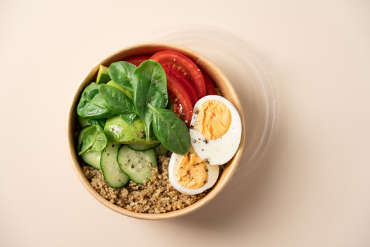 Healthy protein bowl with egg, avocado, spinach, cucumber and quinoa in a cardboard container on a light beige background, nutritious balanced meal concept 