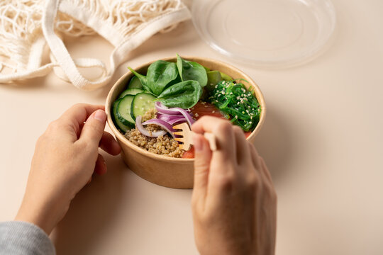 Hands holding a healthy protein bowl with quinoa, smoked salmon, spinach, cucumber, avocado and chuka seaweed salad in a cardboard container on a light beige background, healthy lifestyle concept