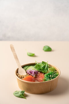 Healthy protein bowl with quinoa, smoked salmon, spinach, cucumber, avocado and chuka seaweed salad in a cardboard container on a light beige background, balanced nutritious meal