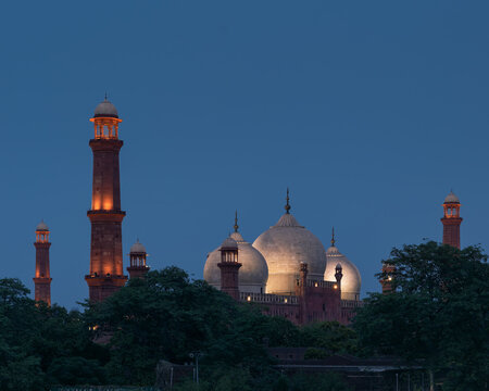 View of the illuminated domes and minarets of the historic Badshahi Mosque at twilight, set against a deep blue sky with lush trees in the foreground in Lahore, Punjab, Pakistan.