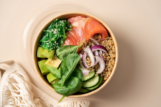 Healthy protein bowl with quinoa, smoked salmon, spinach, cucumber, avocado and chuka seaweed salad in a cardboard container on a light beige background, balanced nutritious meal