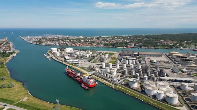 Aerial drone view of red gas tanker ship docked at Ravenna port near chemical refinery plant with gas flare stack burning bright on sunny day in Emilia Romagna Italy 