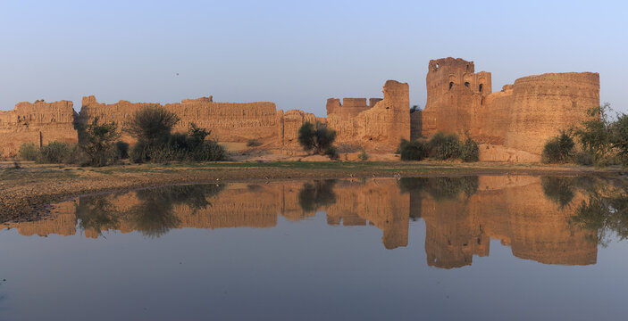 View of the historic Mauj Garh Fort with its ancient mud-brick walls reflected in a calm pool of water under a clear sky in Mauj Garh, Punjab, Pakistan.