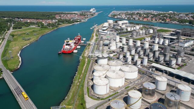 Aerial drone view of red gas tanker ship docked at Ravenna port near chemical refinery plant with gas flare stack burning bright on sunny day in Emilia Romagna Italy 