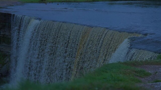 A waterfall fills the frame on an overcast early morning, with flowing water spanning the full width and soft green grass blurred in the foreground.

