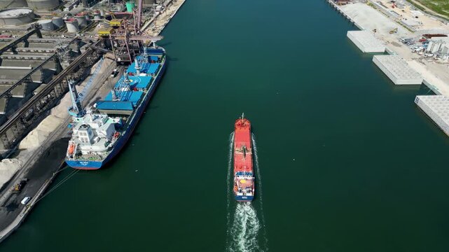 Top down aerial drone view of cargo ship sailing along Ravenna port canal past docked vessel and industrial grain silos in Emilia Romagna, Italy