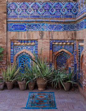 View of the historic Sawi Mosque featuring intricate blue kashi-kari tilework, brick walls, and potted aloe vera plants with a prayer rug in Multan, Punjab, Pakistan.