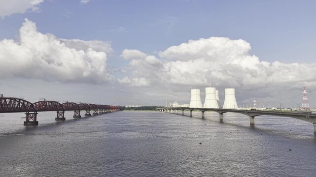 Drone flying forward between Hardinge Bridge and Lalon Shah Bridge over the full Padma River, revealing Rooppur Nuclear Power Plant cooling towers, Green City Rooppur buildings, blue sky, and clouds.