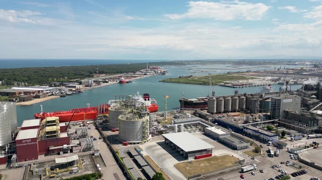 Aerial drone view of red gas tanker ship docked at Ravenna port near chemical refinery plant with gas flare stack burning bright on sunny day in Emilia Romagna Italy 