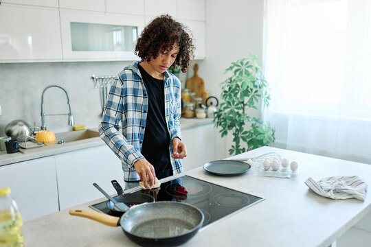 A young person teenager cooks breakfast in a bright modern kitchen, frying an egg in a small skillet on the stovetop. Natural light fills the space and plants and utensils are visible, conveying a rel