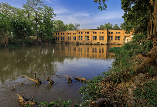 View of Sir Ganga Ram hydro power project with historic brick architecture reflected in calm river water, surrounded by lush trees in Renala Khurd, Punjab, Pakistan.