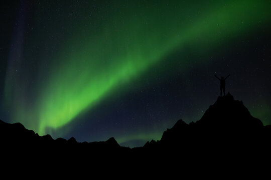 Silhouette of a figure on a peak under a stunning, colorful northern lights sky.